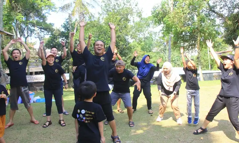 Sekelompok peserta pria, wanita, dan anak-anak tampak ceria mengangkat tangan saat mengikuti sesi fun games luar ruangan dalam acara family gathering di Bogor. Peserta mengenakan kaos seragam hitam dengan logo BoundEx di area lapangan rumput yang asri.