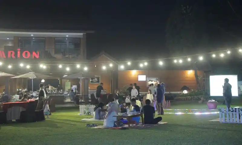 Sekelompok peserta family gathering di Bogor sedang menikmati makan malam lesehan di area rumput terbuka di depan bangunan bertuliskan 'ARION' pada malam hari. Foto menampilkan peserta duduk santai di tikar, dekorasi lampu gantung, dan layar proyeksi di kejauhan.