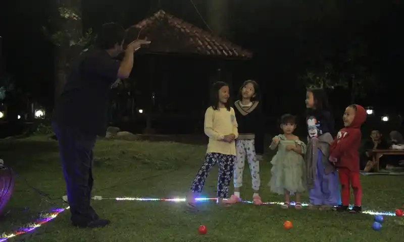 Anak-anak peserta family gathering di Bogor dengan asyik mengikuti sesi fun games interaktif pada malam hari di area BoundEx. Foto menunjukkan instruktur yang sedang memandu permainan di antara dekorasi lampu gantung dan hias.