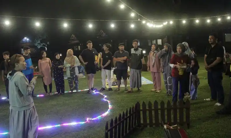 Sekelompok peserta family gathering di Bogor sedang mengikuti sesi ice breaking malam hari di area rumput terbuka dengan dekorasi lampu LED warna-warni dan lampu gantung. Foto menampilkan instruktur BoundEx memberikan pengarahan di depan peserta.