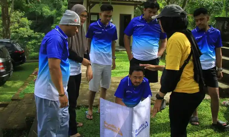 Tim BoundEx Indonesia sedang memberikan instruksi simulasi team building outdoor kepada peserta gathering perusahaan di Bogor. Instruktur (baju kuning) memandu peserta (baju biru) menggunakan alat peraga visual di lapangan rumput yang luas.