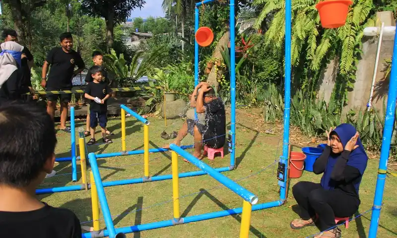 Sekelompok peserta family gathering di Bogor, terdiri dari pria, wanita berhijab, dan anak-anak, sedang mengikuti permainan fun game Mouse Trap versi besar di lapangan rumput asri. Peserta pria di kursi merah tampaknya terkena hukuman air, sementara peserta lain dan penonton di area BoundEx tersenyum dan bereaksi dengan ceria di dekat instalasi pipa biru dan kuning.
