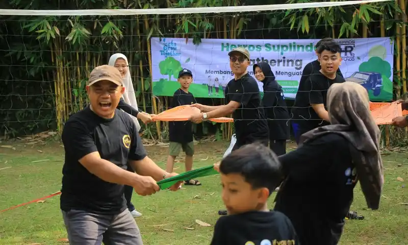 Sekelompok peserta family gathering di Bogor, terdiri dari pria, wanita berhijab, dan anak-anak, sedang bekerja sama melempar kain sarung oranye dan hijau dalam permainan Volley Sarung di area terbuka. Di latar belakang terdapat net voli dan spanduk 'Herros Supindo Gathering' di area asri Puncak.