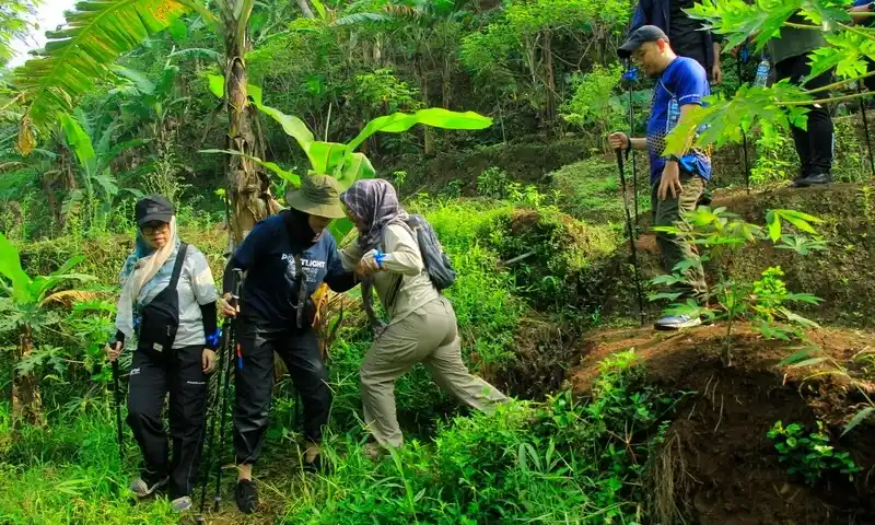 Peserta gathering sedang melakukan trekking di jalur setapak hutan Bogor, saling membantu saat melewati medan tanah yang menanjak.