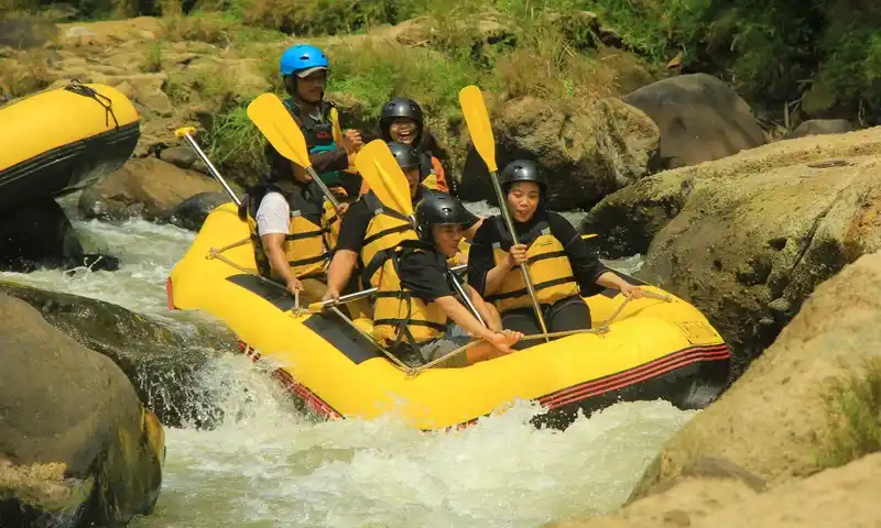 Sekelompok peserta family gathering di Bogor sedang menikmati aktivitas rafting (arung jeram) di sungai berbatu dengan jeram yang cukup deras. Peserta mengenakan jaket pelampung kuning, helm pengaman hitam dan biru, memegang dayung kuning. Di latar belakang terlihat bagian perahu lain.