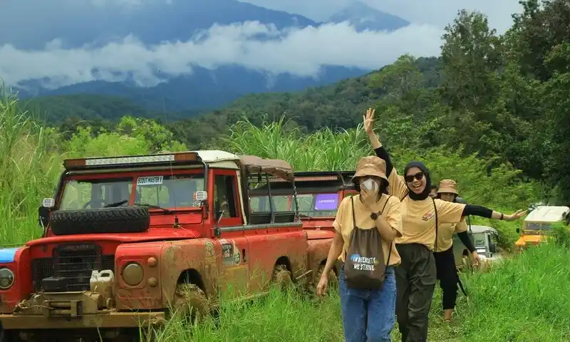 Peserta gathering perusahaan sedang menikmati petualangan offroad di Bogor menggunakan kendaraan Land Rover merah melalui jalur tanah yang menantang. Tampak ekspresi ceria karyawan dengan latar belakang pemandangan pegunungan dan hutan hijau yang dikelola oleh BoundEx Indonesia.