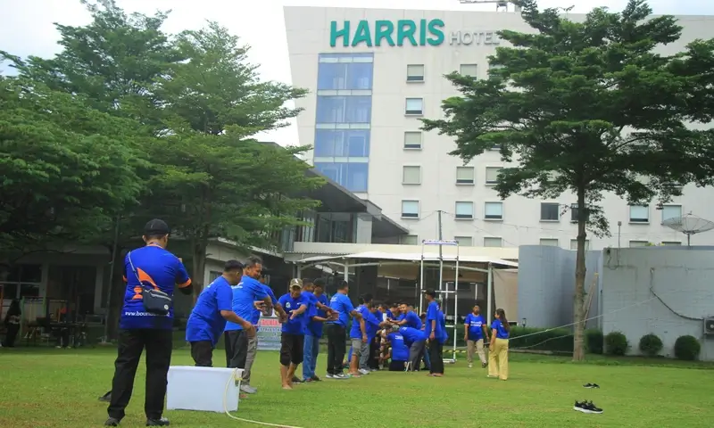 Sekelompok peserta berbaju biru sedang melakukan simulasi kerja sama tim di lapangan rumput Harris Hotel Sentul City. Di latar belakang terlihat gedung hotel putih yang modern dan pepohonan hijau yang rindang dalam rangkaian acara family gathering di Bogor.