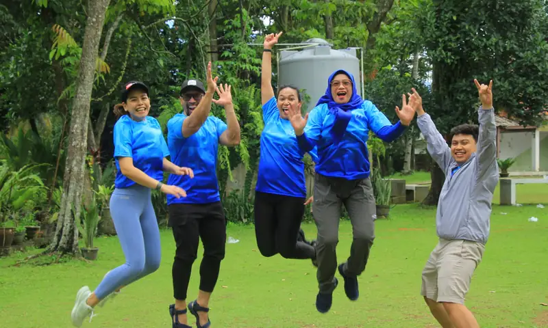 Peserta gathering berkaus biru melompat ceria di area outdoor hijau.