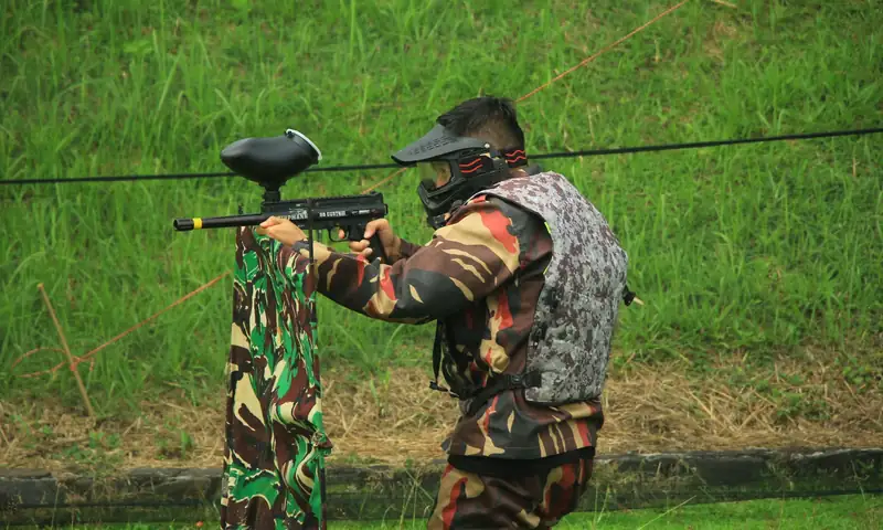 Peserta paintball menggunakan pelindung wajah dan marker dalam aktivitas outbound di area hijau